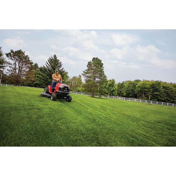 Person operating a riding lawn mower on a green lawn with trees and a white fence in the background.