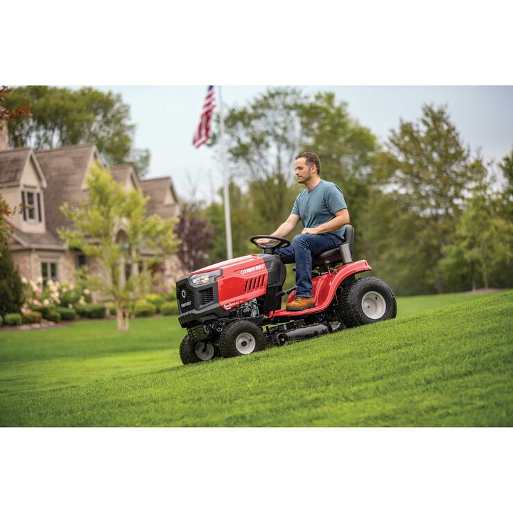 Man operating a red riding lawn mower on a green lawn with a house and trees in the background.